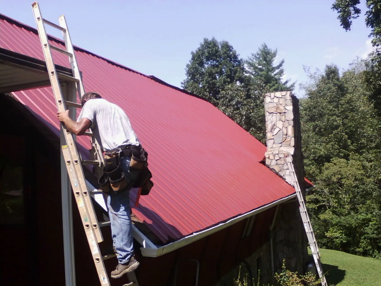 Roofer on a ladder beside a red metal roof and stone chimney in Kenesaw